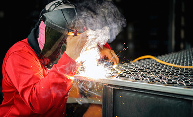 Welder wearing a red long-sleeved shirt and a protective mask is welding a steel grating,...