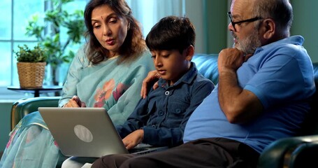 Indian kid with grandparents using laptop computer while sitting on sofa in modern home indoors, elderly grandmother and grandfather teaching child digital learning, family bonding, technology - Powered by Adobe