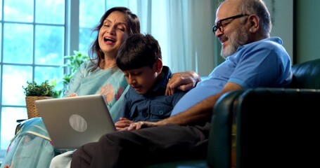 Indian kid attending video call with grandparents using laptop computer while sitting on sofa in modern home indoors, child smiling and waving to grandmother, grandfather, family bonding technology - Powered by Adobe