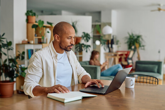 Man using laptop and notebook, working remotely in his living room, while a woman relaxes on the sofa in the background