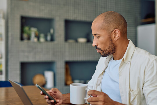 Businessman holding a mug and using his smartphone while working remotely in his modern kitchen - Powered by Adobe