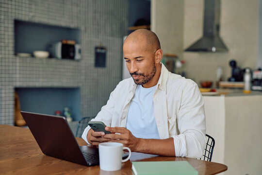 Man working from home using laptop and smartphone, sitting at wooden table with coffee mug and notebook