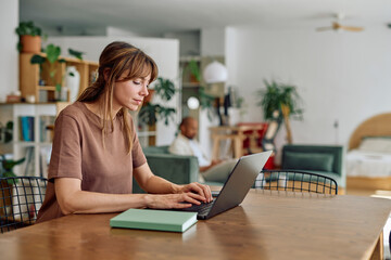 Young woman concentrating while using laptop for working remotely in home office, her partner...