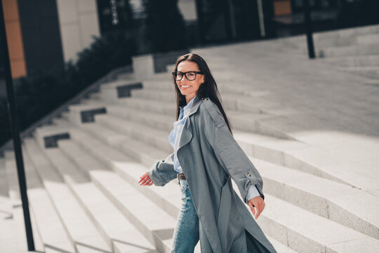 Mature Woman in Grey Trench Coat Walking Outdoors in Modern Urban Setting on a Sunny Day