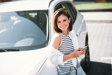 Fototapeta premium Charming young woman smiling in daylight while leaning against car in urban area