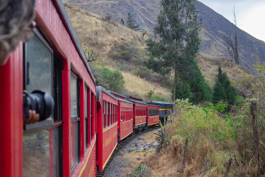 Scenic view of a red train winding through a mountain valley with passengers taking photos, surrounded by trees, rocky terrain, and dramatic Andean landscapes.