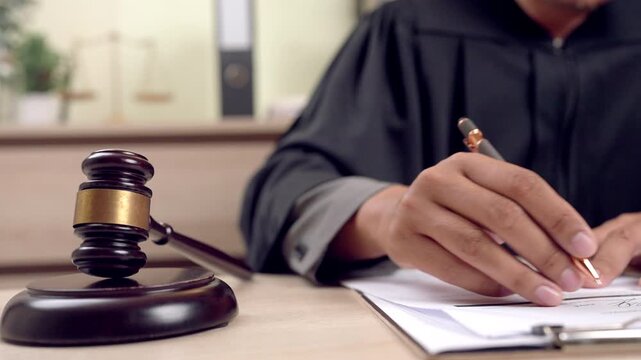 Close-up of judge s hands signing documents beside gavel and justice scale. Concept of law enforcement and decision-making.