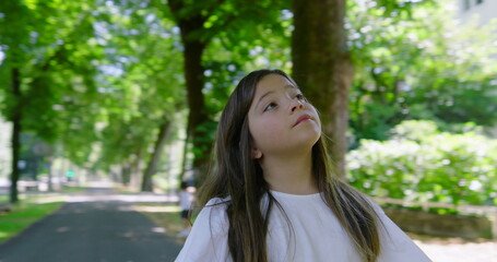 Girl with long hair looking upwards in a tree-lined park, wearing a white blouse, surrounded by...