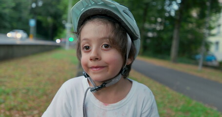 Boy with protective gear sitting on a skateboard by the roadside, helmet on, looking at passing...