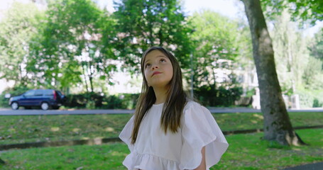 Naklejka premium Girl with long hair looking upwards in a tree-lined park, wearing a white blouse, surrounded by lush greenery and bright sunlight, conveying a sense of wonder and curiosity-SD 480p