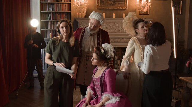 Medium full shot of female movie producer and actors in Victorian period costumes discussing directions in script, technician preparing lighting equipment, makeup artist powdering actress face