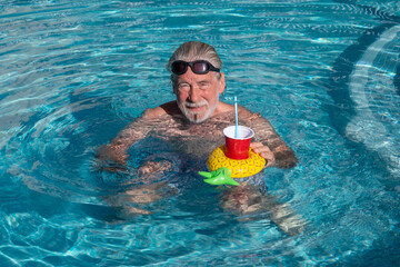 Smiling senior man relaxing  floating at a swimming pool. A floating drink holder create a cheerful summer vacation atmosphere