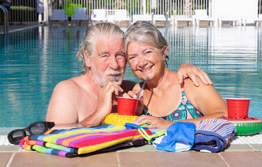 Happy senior couple relaxing together at a swimming pool, smiling and embracing at the poolside....