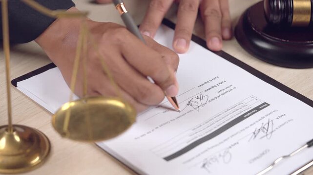 Close-up of hand signing an official legal document with justice scale and judge gavel in the frame. Law and legal concept.