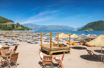 Sandy beach in Icmeler, popular coastal resort in Turkey, with rows of white sunbeds and straw parasols along turquoise shoreline. Icmeler surrounded by green hills and calm sea under blue sky