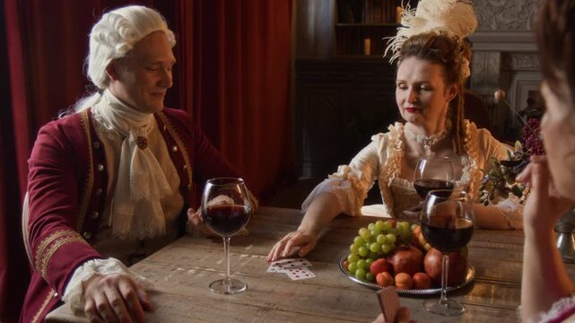 Medium shot of noble Victorian period gentleman in red surcoat and wig and two middle-aged lady friends sitting around table in palace at daytime, enjoying red wine and playing card game
