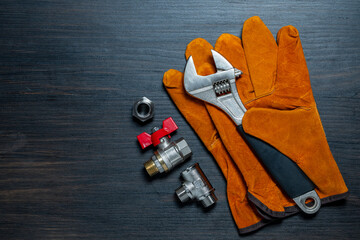 Adjustable wrench, ball valve with red handle, stainless steel tee fittings, leather safety gloves and rubber gaskets on wooden background, top view, copy space, closeup. Plumbing repair concept