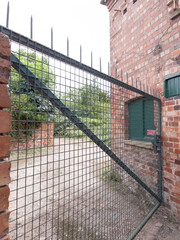 Fototapeta premium A large, industrial metal gate against a red brick building, offering a view into an overgrown, outdoor area