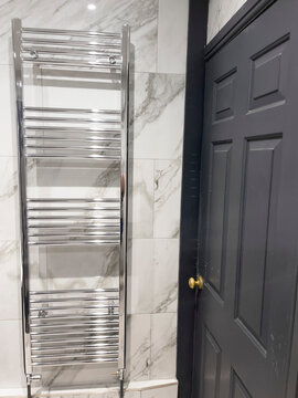 A chrome towel rail with horizontal bars is mounted on a wall of large white marble tiles. The tiles have grey veining and a polished surface. 
