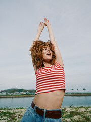 Playful young woman in a red and white striped shirt dances with raised arms by a lake, expressing...