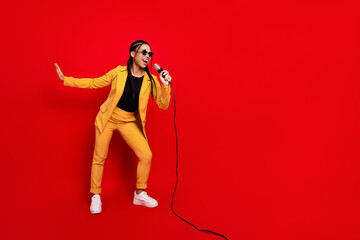 Vibrant young woman in yellow suit holding microphone and singing energetically against a vivid red backdrop