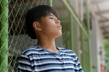 Teen boy standing behind a chain-link fence, looking up seriously and thoughtful. Concept of...