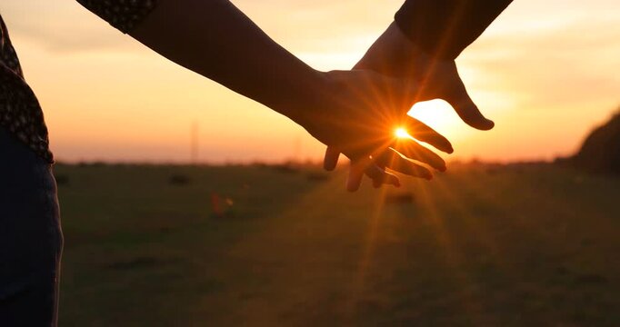 Close-up of the silhouettes of a couple in love holding their hands together, romantic scene at sunset, slow motion. The man and woman have their hands clasped tightly and the golden sun