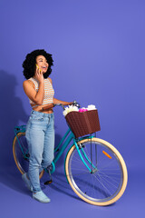 Joyful young woman stands with bicycle beside floral basket, wearing colorful tank top and casual...