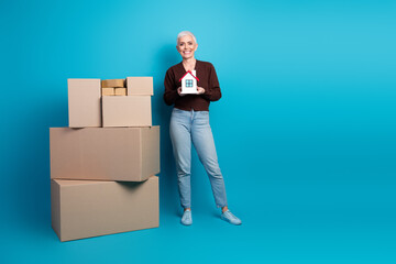 Smiling elderly woman holding a house model near large cardboard boxes on a blue background...