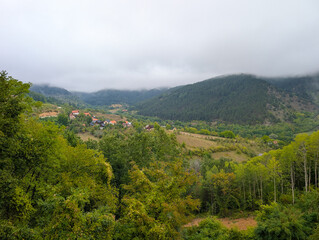 Fototapeta premium Foggy mountains and a valley with small houses and red roofs among green trees. Peaceful rural living and natural beauty in the Balkans.