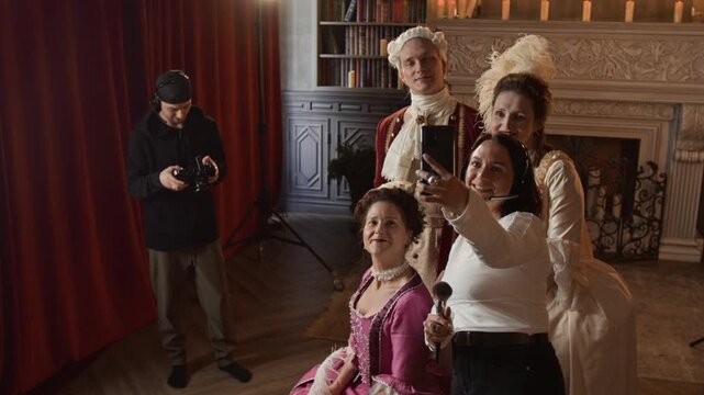 Medium full shot of cheerful female makeup artist taking smartphone selfie with actors in costumes on movie set of historical drama series, all smiling, while cameraman is checking filmed footage