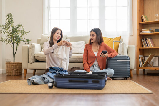 Young asian woman and friend preparing luggage packing clothes in suitcases for travel at home, planning holiday travel, content creator and travel blogger, friendship moments lifestyle vacation trip