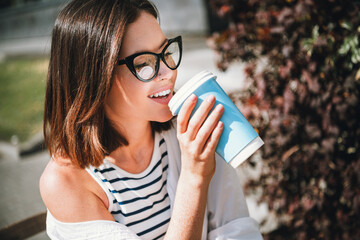 Young woman enjoying coffee outdoors on a sunny day, wearing stylish casual clothes, embracing the...