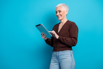 Cheerful mature woman enjoying leisure time and using a digital tablet against a vibrant blue...