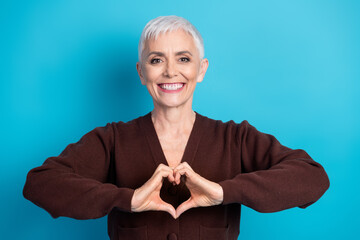 Confident mature woman with short grey hair making a heart gesture on a blue background, showcasing positivity