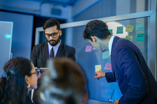 A diverse team of project managers uses a whiteboard and sticky notes for a brainstorming session. They are collaborating on a new business strategy in their corporate office.