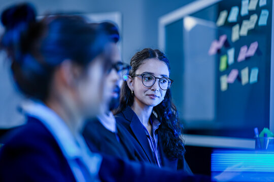 An agile development team participates in a sprint retrospective meeting. A female developer contemplates the user stories on the kanban board visible in the background.
