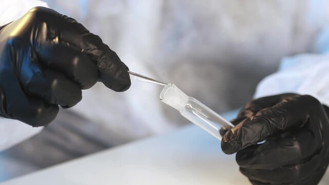 A forensic scientist wearing a protective suit and gloves removes a human hair from a test tube. A forensic scientist removes evidence from a glass test tube in a lab for a DNA test.