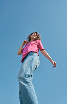 Beautiful carefree young woman with curly hair in pink t-shirt and jeans smiling and looking aside on blue sky background low angle shot.