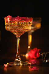 Two elegant champagne glasses with sparkling wine and rose petals, set on a wooden floor with candlelight and a dark, intimate background.