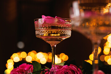 A close-up of a delicate champagne coupe filled with golden bubbles and pink rose petals, surrounded by fresh roses and illuminated by dreamy warm lights.
