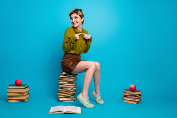 Clever young woman in trendy clothing sits on book against blue background, showcasing vibrant...