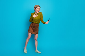 Young stylish woman holding a magnifying glass amazed against a blue background portraying fashionable casual lifestyle