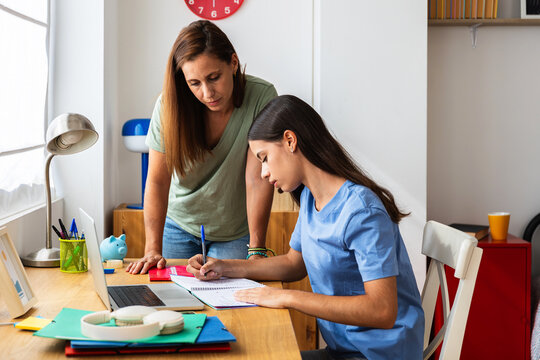 Mid adult mother helping teenage daughter with her homework while studying on laptop in her bedroom. Back to school and education lifestyle concept