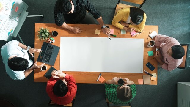 Top down aerial view of diverse business team designing and planning project while skilled manager walking and joining meeting at meeting table with sticky notes and laptop at boardroom. Convocation.