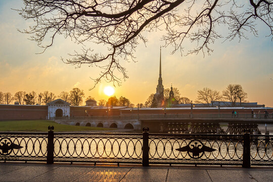 Ornate cast iron fence with double-headed eagle emblem in sunset backlight, with Peter and Paul Fortress silhouette across Neva River. Saint Petersburg, Russia.