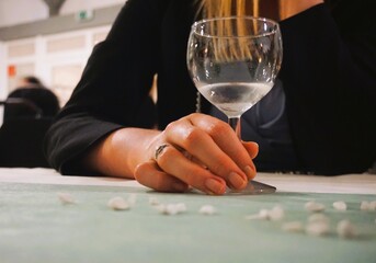 Junge schlanke attraktive Frau mit blonden langen Haaren und schwarzem Oberteil sitzt vor Tisch und umfasst ein leeres Weinglas auf Feier am Abend im Winter