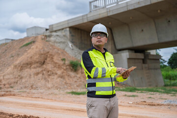 Construction engineer wearing safety gear and holding a tablet inspects bridge development site....