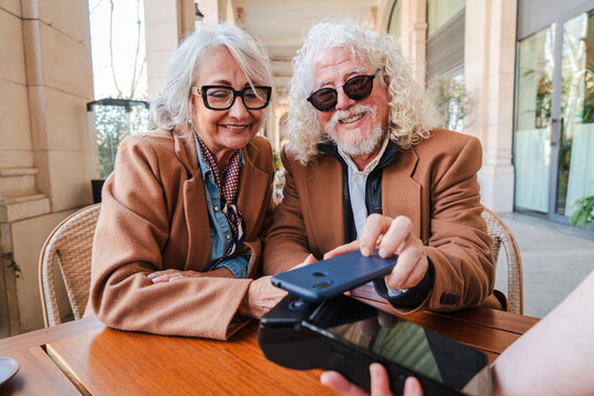 A happy senior couple actively participating in a seamless payment process using their smartphone at a dining table, illustrating their enjoyment of technology and shared moments while managing