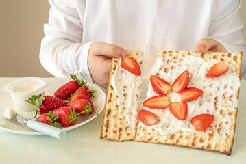 Matzah covered with creamy cheese and sliced strawberries in the hands of a child for tasty breakfast during Jewish holiday Pesach or Passover celebration.